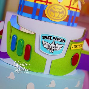 May include: A multi-tiered cake featuring designs inspired by the movie Toy Story. The top tier is decorated to resemble Woody's outfit, while the middle tier is Buzz Lightyear's space suit. The bottom tier is light blue with white clouds.