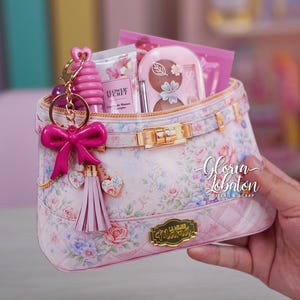 May include: A pink floral patterned handbag with gold hardware, filled with beauty products. A pink bow and tassel keychain hangs from the zipper. The bag features the text "La Mejor Mama" and "Gloria Jobaton".