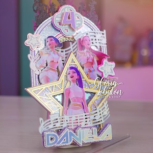 May include: A decorative cake topper featuring three cartoon figures, a star, and musical notes. The topper has the number 4 and the name "DANIELA" in glittery letters. The design includes pink, purple, and silver accents.