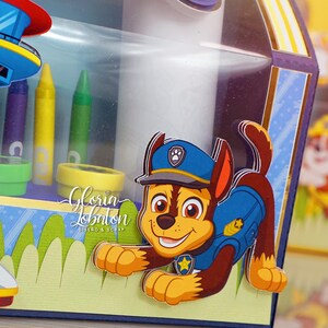 Pode incluir: Uma caixa de cart&atilde;o decorada com um personagem da Patrulha Canina, Chase, em seu uniforme de policial. A caixa tem uma janela de pl&aacute;stico transparente e &eacute; decorada com um esquema de cores azul, amarelo e verde. O texto "Gloria Lobaton Dise&ntilde;o & Scrap" &eacute; vis&iacute;vel na caixa.