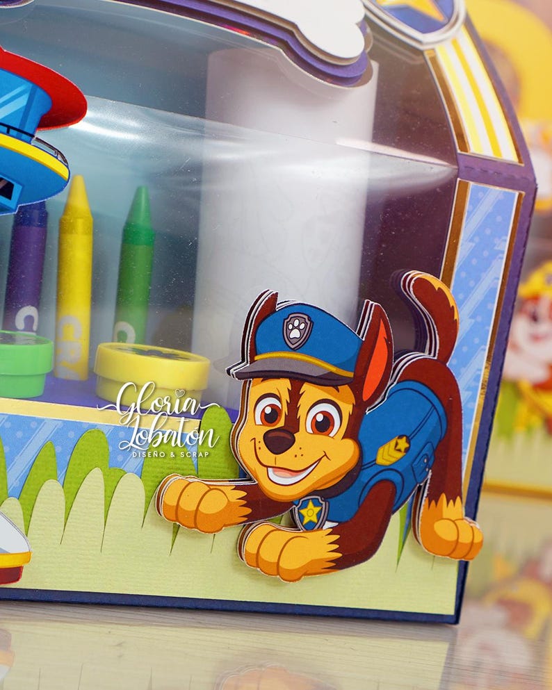 Pode incluir: Uma caixa de cart&atilde;o decorada com um personagem da Patrulha Canina, Chase, em seu uniforme de policial. A caixa tem uma janela de pl&aacute;stico transparente e &eacute; decorada com um esquema de cores azul, amarelo e verde. O texto "Gloria Lobaton Dise&ntilde;o & Scrap" &eacute; vis&iacute;vel na caixa.