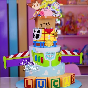 May include: A multi-tiered cake decorated with Toy Story characters. The cake features Woody, Buzz Lightyear, and other characters. The bottom tier has wooden blocks spelling out the name "LUCAS". The cake has a blue base and is decorated with clouds.
