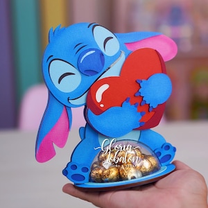 以下が含まれることがあります： 赤いハートを持った青い漫画のキャラクター。キャラクターは、包装されたチョコレートが入った透明なドームが付いた青いベースにいます。デザインには「Gloria Lobaton, Diseño Scrap」という文字が含まれています。