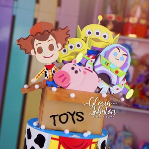 May include: A colorful cake featuring characters from the animated film Toy Story. The cake includes a cardboard box labeled "TOYS" with Woody, Buzz Lightyear, and other characters. The cake is decorated with a sheriff's badge and a cowhide pattern.