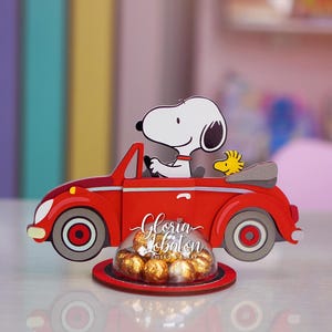 Könnte beinhalten: Ein roter, Snoopy-förmiger Bonbonhalter in Form eines Autos mit Snoopy am Steuer und Woodstock als Beifahrer. Er enthält goldverpackte Pralinen unter einer transparenten Kuppel. Auf dem Sockel steht "Gloria Lobaton".