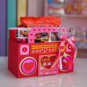 Puede incluir: Una caja de regalo roja decorada con corazones y un diseño de cinta de cassette, llena de snacks y bebidas. La caja tiene un asa con recortes de corazón y un lazo decorativo. El texto "LOVE MIX" es visible.