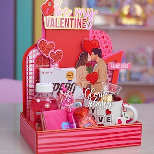 Puede incluir: Una cesta de regalo de San Valentín con un tema rojo y rosa. Incluye una taza, una botella de líquido rosa, una cartera rosa y una caja de caramelos. Las palabras "Be My Valentine" se muestran en la parte superior.