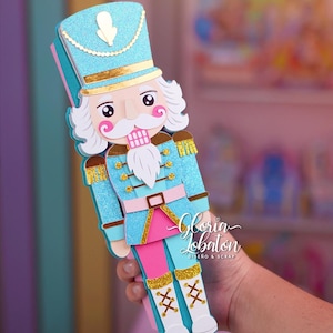 Puede incluir: Una figura decorativa de cascanueces azul y rosa brillante. La figura tiene un sombrero azul con ribete dorado, barba y bigote blancos y mejillas rosadas. El cascanueces sostiene un objeto rosa y azul. El texto "Gloria Lobaton" es visible.