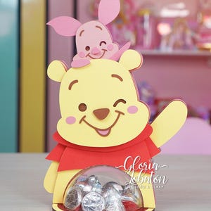 May include: A yellow and red Winnie the Pooh character cutout with a pink Piglet character cutout on top. The cutout has a clear plastic container in the front for holding small candies. The text "Gloria Lobaton Diseño & Scrap" is printed on the bottom of the cutout.
