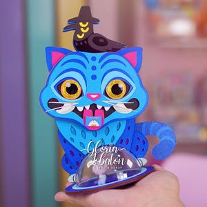 Peut inclure: Une œuvre d'art en papier superposé fantaisiste représentant un chat bleu avec de grands yeux jaunes et un oiseau portant un chapeau. Le chat a des accents roses et une queue rayée. Le texte "Gloria Lobaton" et "DISENO & SCRAP" est inclus.