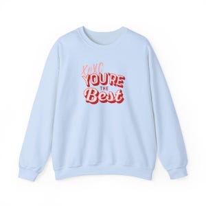 Puede incluir: Sudadera azul claro con la frase "XOXO You're the Best" en rosa y rojo. El texto está en una fuente audaz y juguetona. La sudadera es de cuello redondo y está hecha de un material suave.