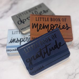 May include: Three small, hardcover journals with different colored covers. The covers are blue, brown, and gray. Each journal has a black inscription on the front. The blue journal says "Little Book of Gratitude", the brown journal says "Little Book of Memories", and the gray journal says "Little Book of Doodles". All three journals have the word "inscribble" printed on the bottom of the cover.