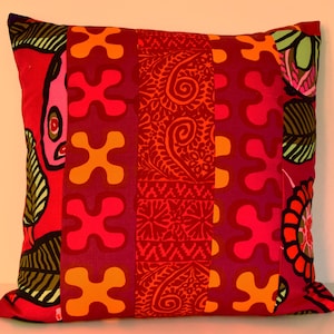 May include: A square throw pillow featuring a patchwork design of vertical fabric strips. The pattern includes vibrant red, orange, and magenta sections with repeating abstract shapes, floral motifs, and intricate paisley-like swirls in olive green.