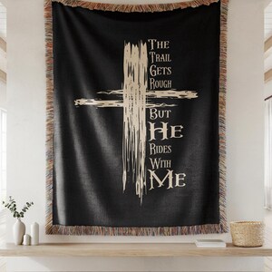 May include: Black woven throw blanket featuring a cream-coloured cross design and the text "THE TRAIL GETS ROUGH BUT HE RIDES WITH ME." The blanket has a fringed edge and is displayed on a wooden shelf.