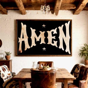 May include: A framed black sign with the word "AMEN" in large, ornate, beige letters. Two small white stars flank the text. The sign hangs above a wooden dining table with leather chairs, creating a rustic, farmhouse aesthetic.