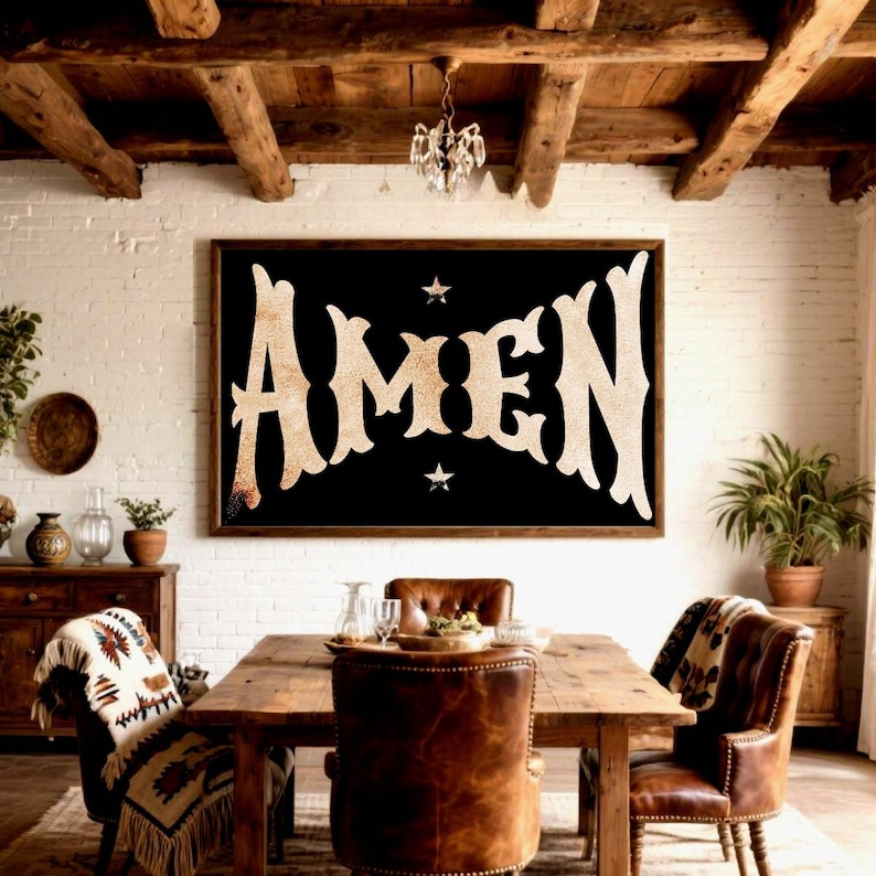 May include: A framed black sign with the word "AMEN" in large, ornate, beige letters. Two small white stars flank the text. The sign hangs above a wooden dining table with leather chairs, creating a rustic, farmhouse aesthetic.
