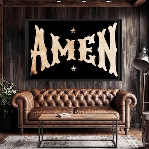 May include: A framed black wall art piece with the word "AMEN" in a gold, textured font. Two small stars flank the text. The artwork hangs above a brown leather Chesterfield sofa and a wooden coffee table.