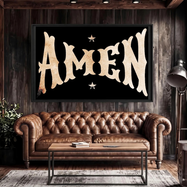 May include: A framed black wall art piece with the word "AMEN" in a gold, textured font. Two small stars flank the text. The artwork hangs above a brown leather Chesterfield sofa and a wooden coffee table.