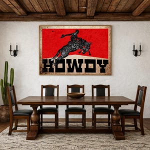 May include: A framed wall art print featuring a rodeo scene with a cowboy riding a bucking bronco against a red background. The word "HOWDY" is displayed in bold, black letters. The print is displayed above a wooden dining table.