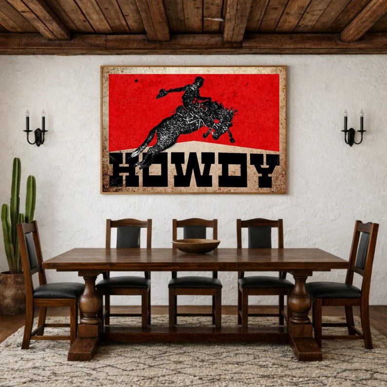 May include: A framed wall art print featuring a rodeo scene with a cowboy riding a bucking bronco against a red background. The word "HOWDY" is displayed in bold, black letters. The print is displayed above a wooden dining table.