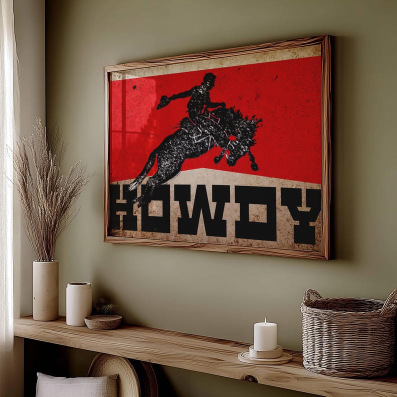 May include: Framed art print featuring a silhouette of a rodeo rider on a bucking bronco against a red background. The word "HOWDY" is displayed in bold, black letters. The artwork is encased in a wooden frame, adding a rustic touch to the western-themed design.