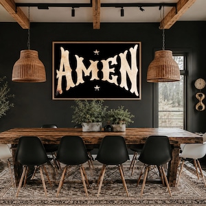 May include: A large framed sign with the word "AMEN" in a vintage font, set against a black background with two small stars. The sign hangs above a wooden dining table with black chairs, creating a rustic and spiritual aesthetic.