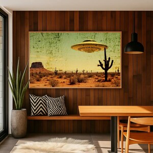May include: A framed vintage-style print depicts a desert landscape with a hovering UFO. The artwork features a yellow and green sky, a brown desert floor, and several cacti. The print has a distressed, aged appearance, adding to its retro aesthetic.