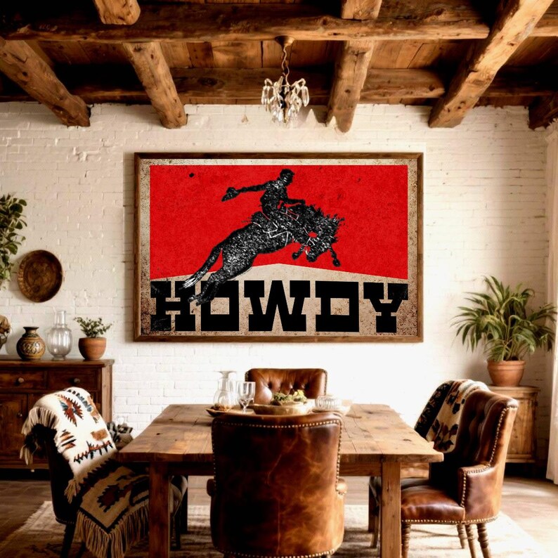 May include: A framed wall art print featuring a rodeo cowboy silhouetted against a red background. The word "HOWDY" is displayed in bold, black letters. The artwork hangs above a wooden dining table with leather chairs, creating a rustic, Western-themed interior.