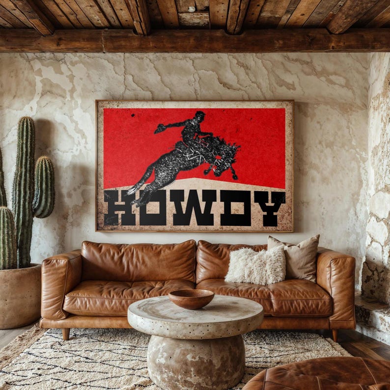 May include: A living room with a brown leather sofa, a round concrete coffee table, and a large framed artwork. The artwork features a black silhouette of a rodeo rider on a bucking bronco against a red background with the word "HOWDY".