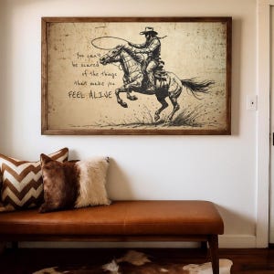 May include: A rectangular framed wall art print featuring a black ink sketch of a cowboy riding a galloping horse while swinging a lasso. The text reads: You can't be scared of the things that make you FEEL ALIVE. The background is aged parchment.