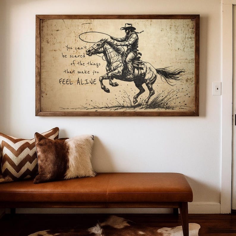 May include: A rectangular framed wall art print featuring a black ink sketch of a cowboy riding a galloping horse while swinging a lasso. The text reads: You can't be scared of the things that make you FEEL ALIVE. The background is aged parchment.
