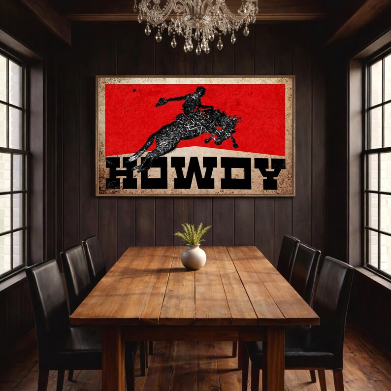 May include: A framed print featuring a rodeo cowboy on a bucking bronco against a red background. The word "HOWDY" is displayed in bold, black letters. The artwork is displayed above a wooden dining table with black chairs.