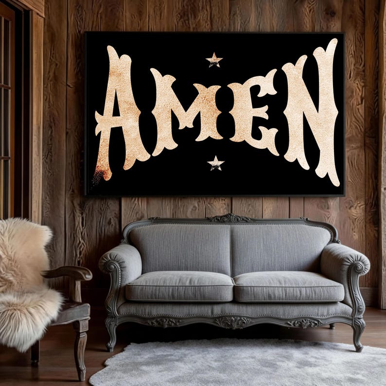 May include: A black framed wall art piece with the word "AMEN" in a vintage style font. The letters are a light tan color with a textured appearance. Two small star shapes are above and below the word. The art is displayed above a gray sofa.