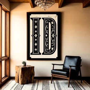 Könnte beinhalten: Ein gerahmtes Wandkunstwerk mit einem großen, stilisierten Buchstaben "D" in Schwarz auf einem cremefarbenen Hintergrund. Der Buchstabe ist mit Zierelementen wie Rauten und Sternen verziert. Das Kunstwerk wird in einem Raum mit einem schwarzen Ledersessel und einem hölzernen Beistelltisch präsentiert.