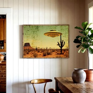 May include: A framed print depicting a desert landscape with a yellow flying saucer hovering above a cactus. The artwork has a vintage, distressed look, with a color palette of yellow, green, and brown. The print is mounted on a wooden frame.