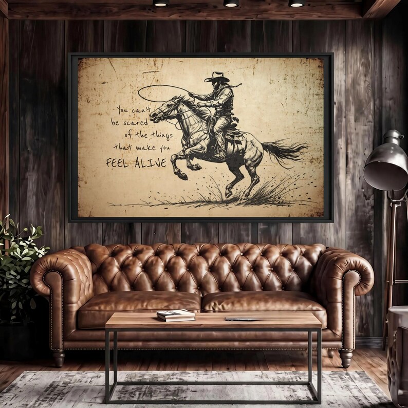 May include: A framed wall art print featuring a black ink sketch of a cowboy riding a galloping horse while holding a lasso. The background is a textured, aged beige paper. The text reads: You can't be scared of the things that make you FEEL ALIVE.