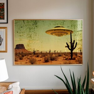 May include: A framed print depicting a desert landscape with a yellow flying saucer hovering above a cactus. The artwork has a vintage, distressed aesthetic, with a muted color palette of tan, green, and yellow. The scene includes cacti, shrubs, and a mesa.