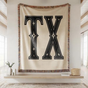 May include: A beige throw blanket with the letters "TX" in large, black, decorative font. The blanket has a fringed edge with a mix of colors. The blanket is hanging on a white wall.