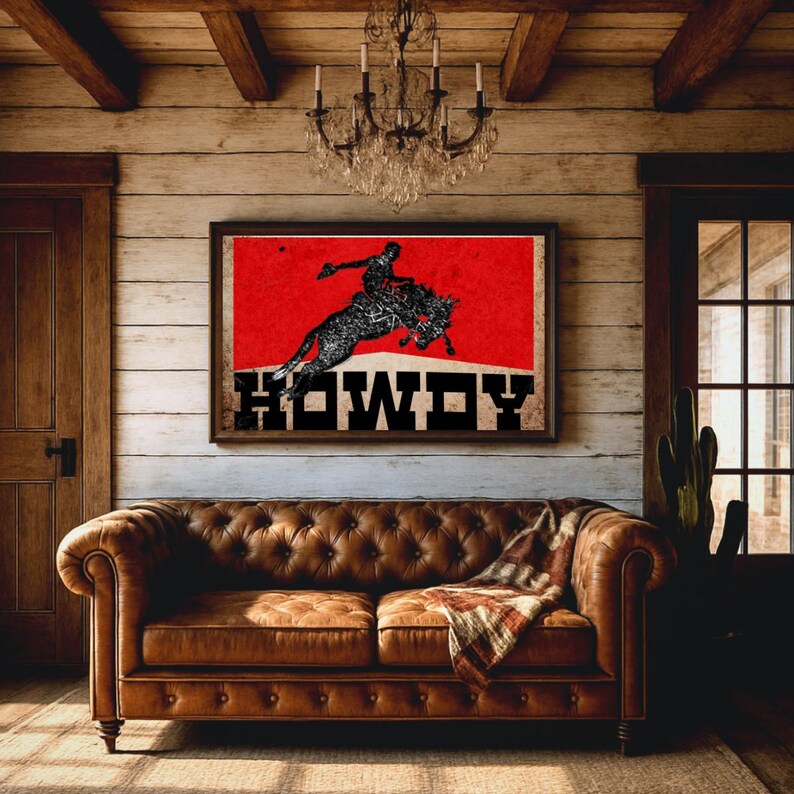 May include: A framed artwork featuring a cowboy riding a bucking bronco against a red background, with the word "HOWDY" in bold letters. The artwork hangs above a brown leather sofa in a rustic-themed room.