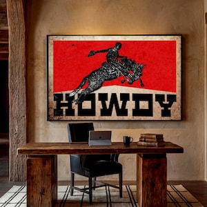 May include: A framed art print featuring a cowboy riding a bucking bronco against a red background. The word "HOWDY" is displayed in bold, black letters. The artwork hangs above a wooden desk with a laptop and books.