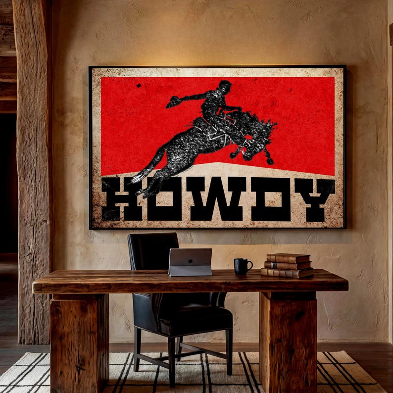 May include: A framed art print featuring a cowboy riding a bucking bronco against a red background. The word "HOWDY" is displayed in bold, black letters. The artwork hangs above a wooden desk with a laptop and books.