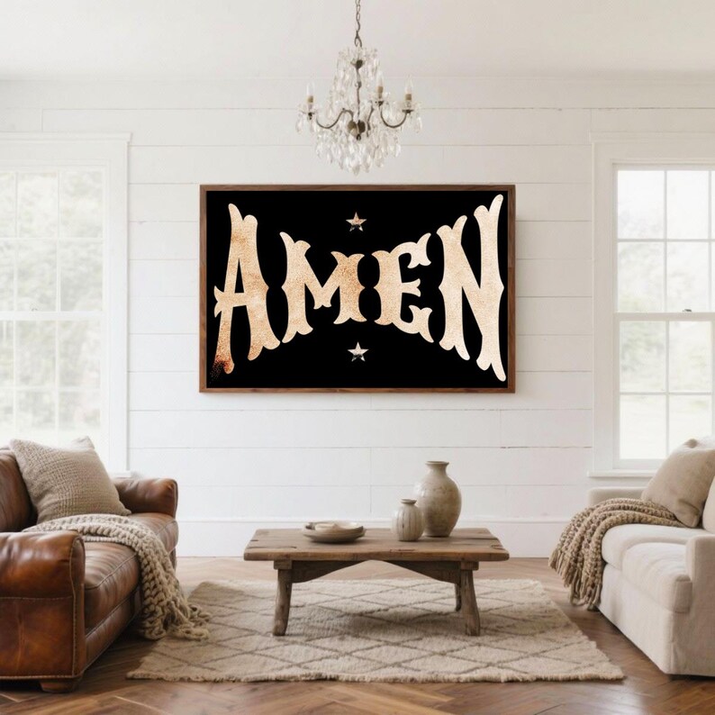 May include: A framed wall art piece with the word "AMEN" in a vintage, distressed font. The text is in a light tan color against a black background, with small star accents. The artwork is displayed in a living room setting.