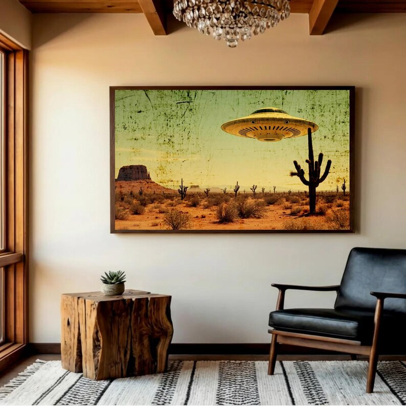 May include: A framed print depicts a desert landscape with a yellow UFO hovering above a cactus. The artwork has a vintage, distressed look, with a color palette of tan, yellow, and green. The print is displayed in a room with a wooden side table and a black leather chair.