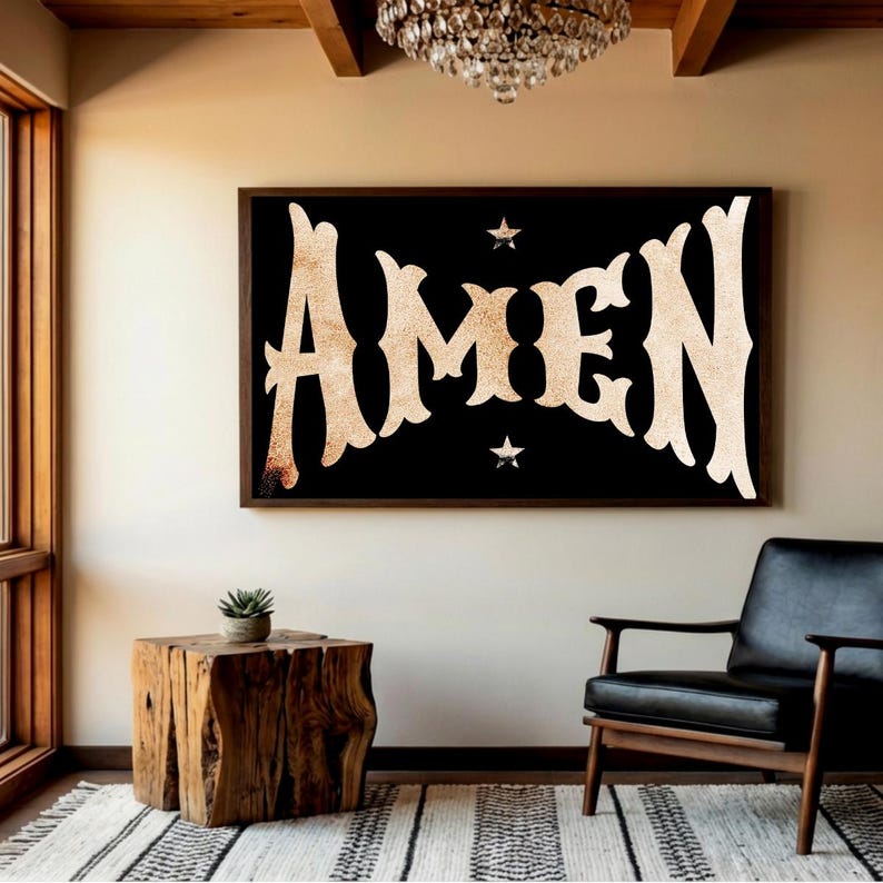 May include: A framed wall art piece with the word "AMEN" in a vintage, textured font against a black background. Two small star shapes are above and below the word. The artwork is displayed in a room with a wooden side table and a black leather chair.
