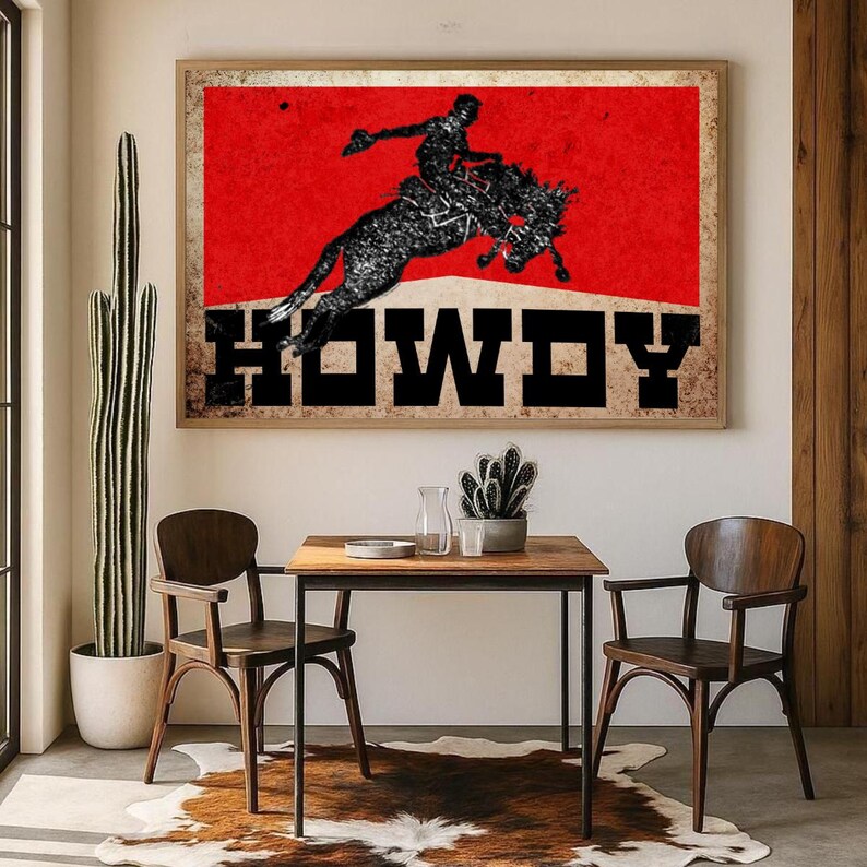May include: Framed wall art featuring a rodeo cowboy silhouette on a bucking bronco against a red background. The word "HOWDY" is displayed in bold, black letters. The artwork is displayed above a small wooden table and chairs.