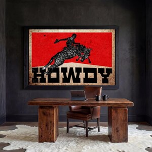 May include: A framed vintage-style print featuring a rodeo scene with a cowboy riding a bucking bronco against a red background. The word "HOWDY" is displayed in bold letters. The print hangs above a wooden desk.