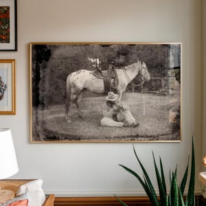 May include: Black and white framed print of a cowboy and horse. The cowboy is seated on the grass, and the horse is standing nearby. The horse has a saddle and bridle. The print is in a gold frame.
