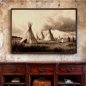 May include: Sepia-toned framed print depicting a Native American village scene. Several teepees stand in a grassy field under a cloudy sky. Two figures stand near a small fire. The artwork is displayed above a dark wood cabinet.