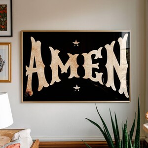 May include: A framed art print with the word "AMEN" in a gold, textured font against a black background. Two small gold stars are above and below the word. The frame is gold.