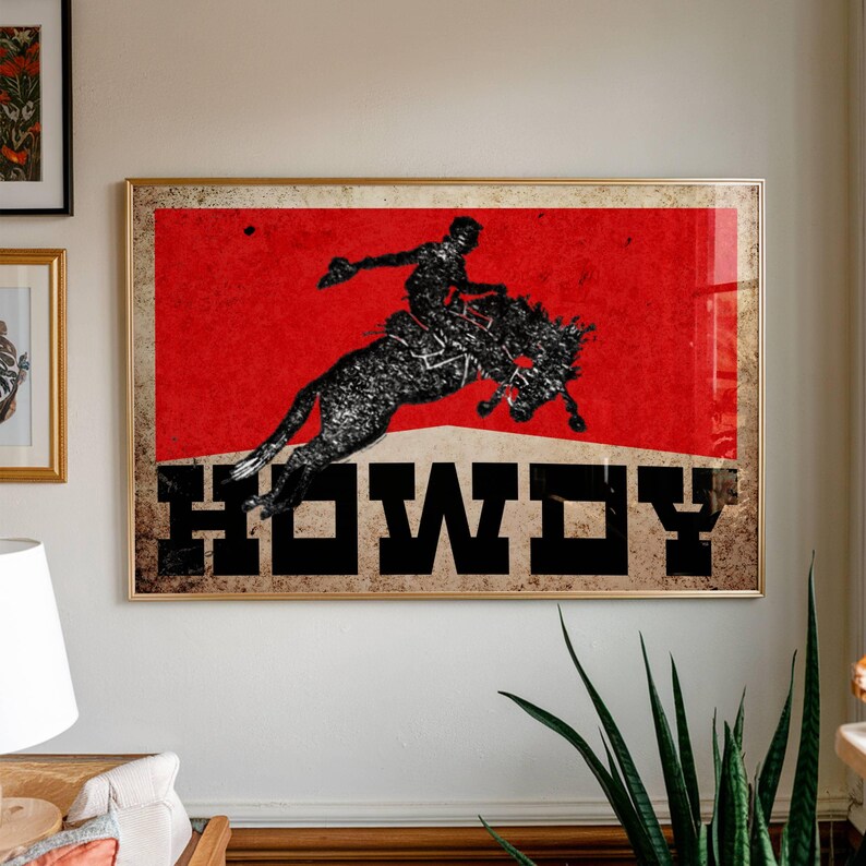 May include: A framed art print featuring a silhouette of a rodeo cowboy riding a bucking bronco against a red background. The word "HOWDY" is printed in bold black letters at the bottom of the image. The artwork has a vintage, distressed look.
